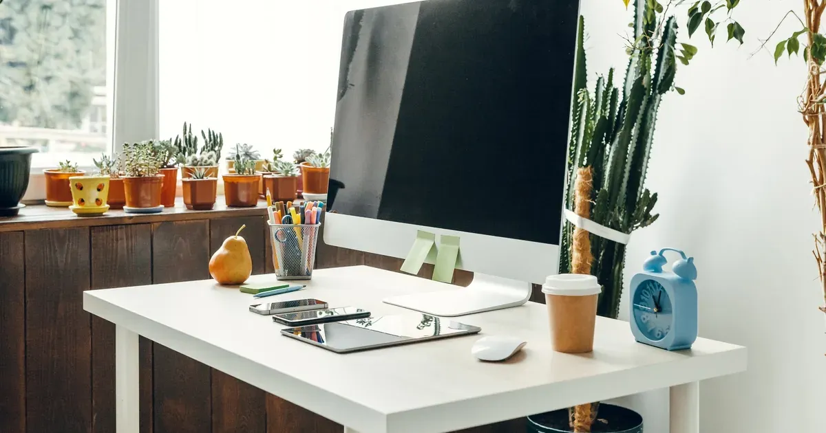 A tidy corner home office with a wooden desk, monitor at eye level, a small plant, and organized cables — natural light from a side window.
