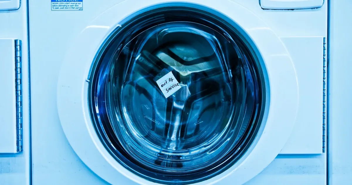 Close-up of a hand scrubbing the rubber door gasket of a front-load washing machine with a toothbrush, vinegar solution visible on the dark rubber folds. Bright laundry room, natural light.
