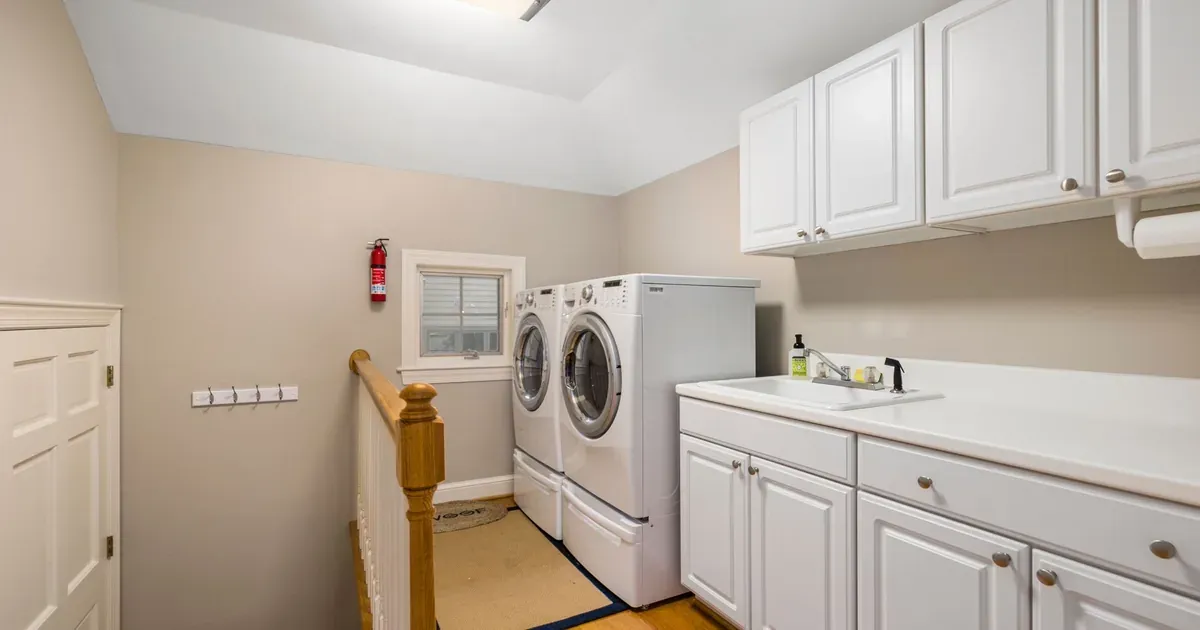 A tidy, compact laundry room with stacked front-loading machines, floating shelves above holding matching white baskets, and a fold-down ironing board mounted to the wall. Natural light, clean lines.