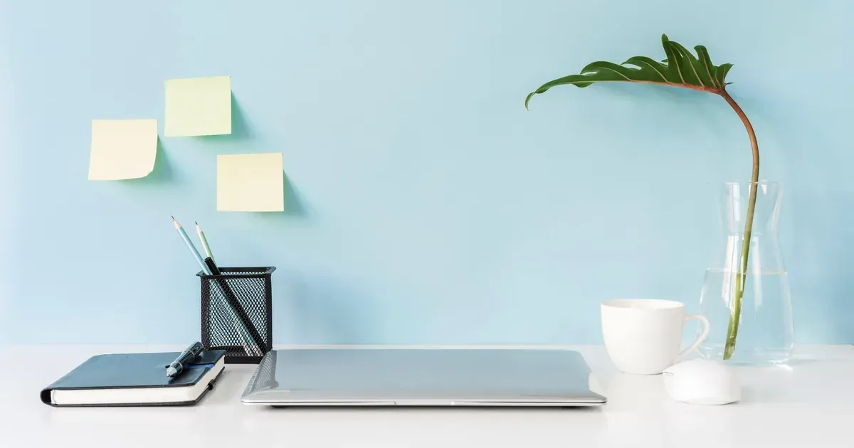 A clean, minimal corner desk setup with a monitor, notebook, and small plant against a white wall, photographed in warm natural light. Conveys calm productivity without clutter.