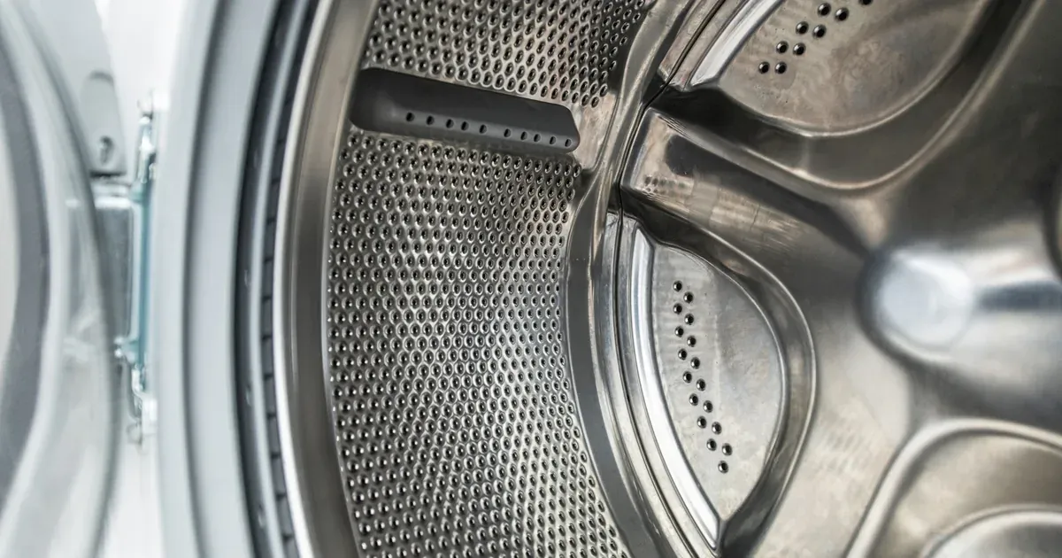 A sparkling clean front-load washing machine drum interior, door open, bright laundry room in background. Before/after contrast implied by the pristine white drum.