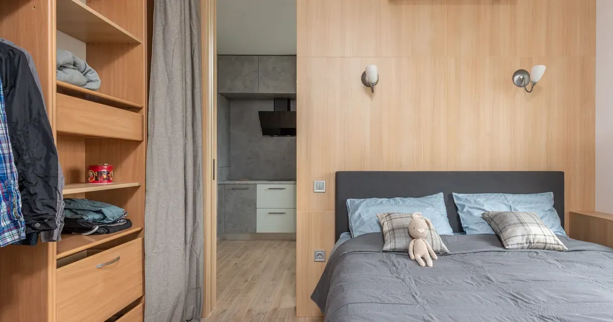 A tidy, well-organized small bedroom with floating shelves, a platform bed with storage drawers, and clear surfaces. Natural light, neutral tones, real-home feel.