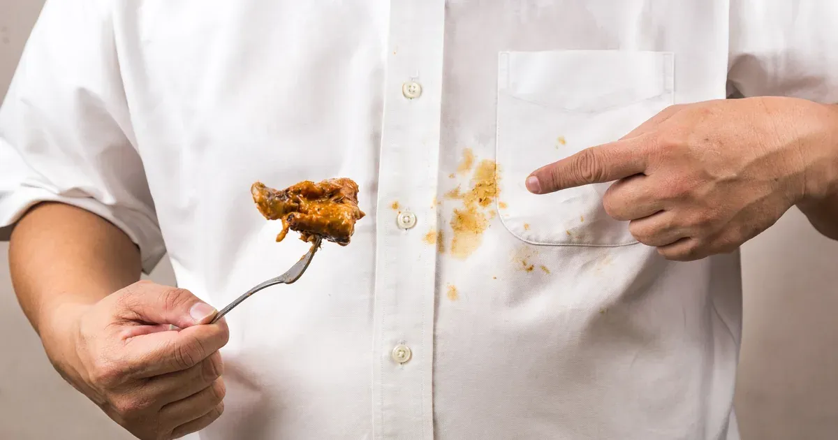 A white cotton shirt with a visible oil stain, a mound of baking soda poured over it, and a small dish soap bottle nearby on a clean countertop. Natural light, close-up shot.