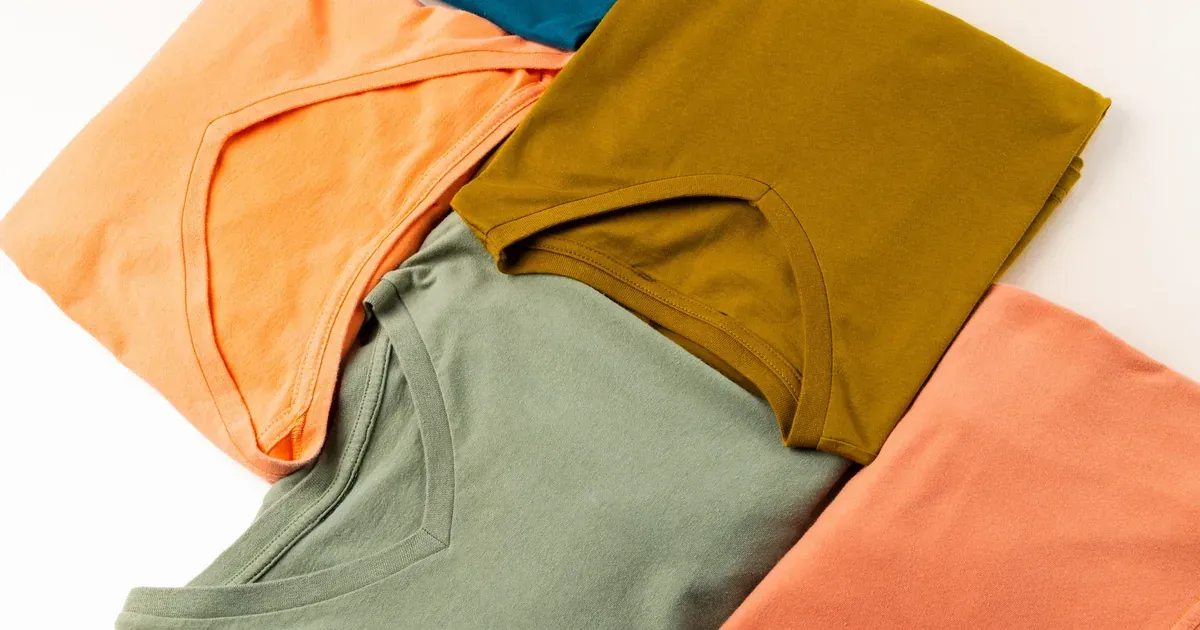 Overhead shot of t-shirts folded and standing upright in a row inside an open drawer, hands mid-fold on a bed nearby.