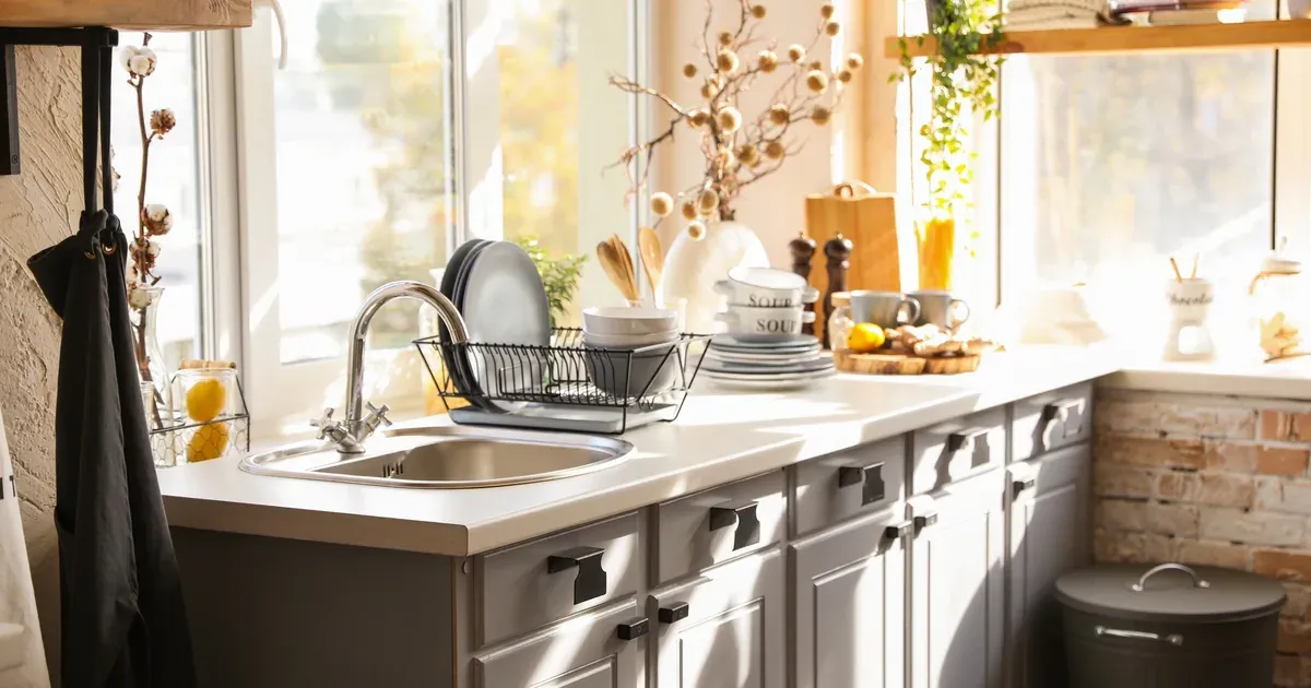 A small, organized kitchen cabinet showing a riser shelf with stacked plates and mugs, sunlight coming through a nearby window.